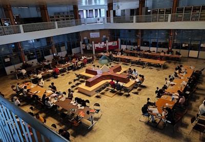 A spacious library interior with multiple long tables arranged in a square pattern, occupied by numerous people who appear to be studying or reading. The library features high ceilings, and bookshelves are visible along the upper level. The overall atmosphere suggests a focused academic environment.