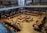 A spacious library interior with multiple long tables arranged in a square pattern, occupied by numerous people who appear to be studying or reading. The library features high ceilings, and bookshelves are visible along the upper level. The overall atmosphere suggests a focused academic environment.
