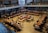 A spacious library interior with multiple long tables arranged in a square pattern, occupied by numerous people who appear to be studying or reading. The library features high ceilings, and bookshelves are visible along the upper level. The overall atmosphere suggests a focused academic environment.