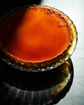 A caramel flan sits in a glass dish, illuminated by a soft light that highlights its glossy, smooth surface. The rich amber color of the caramel contrasts with the darker shadows cast on the surrounding surface.