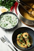 A richly garnished meal featuring two chicken drumsticks in a creamy sauce served in a dark bowl, topped with fresh mint leaves. To the right, a pot contains more of the chicken dish, placed on a wooden board. A bowl of salad with cucumbers and herbs sits on the side. In the background, fresh cilantro, red chili peppers, and a lemon wedge add a touch of color to the scene. A fork and knife are positioned on the left side of the table.