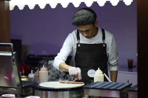 A person wearing a beret and apron is focused on cooking at a food stall or kitchen setup. The individual is using a spatula to cook on a round griddle. Condiment bottles are neatly arranged on the counter, and there is some steam rising from the cooking surface.