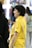 A group of people are gathered in what seems to be an indoor setting, possibly an art gallery or exhibition. A person in a bright yellow shirt is prominently featured, smiling and appearing engaged in conversation. Other individuals are partially visible, dressed in casual attire, some holding items like a drink bottle.