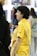 A group of people are gathered in what seems to be an indoor setting, possibly an art gallery or exhibition. A person in a bright yellow shirt is prominently featured, smiling and appearing engaged in conversation. Other individuals are partially visible, dressed in casual attire, some holding items like a drink bottle.