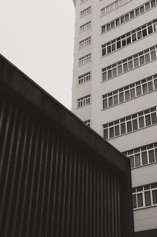A monochromatic photo of an urban setting featuring a modern high-rise building with numerous rectangular windows and a dark structure with vertical lines in the foreground taken from a low angle.