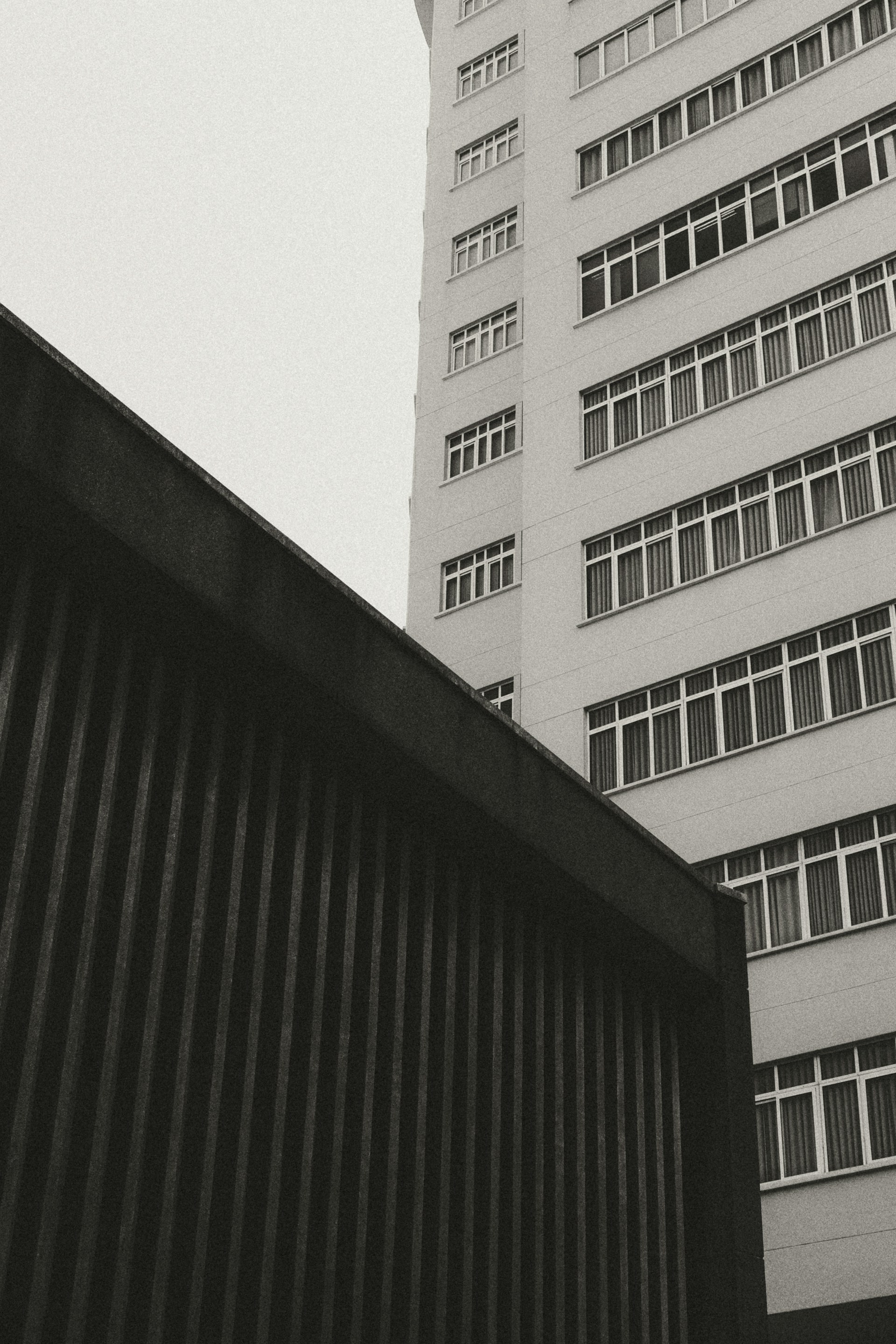 A monochromatic photo of an urban setting featuring a modern high-rise building with numerous rectangular windows and a dark structure with vertical lines in the foreground taken from a low angle.