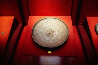 A decorative, round metal object with intricate carvings is displayed against a deep red background. The center features an engraved scene with figures, surrounded by ornate patterns.