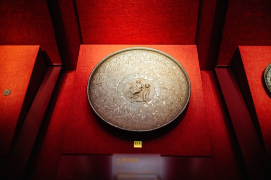 A decorative, round metal object with intricate carvings is displayed against a deep red background. The center features an engraved scene with figures, surrounded by ornate patterns.