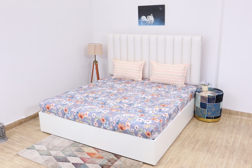 A neatly arranged bedroom features a white upholstered bed with a floral-patterned bedspread and two striped pillows. Next to the bed is a round, multi-colored side table with a small plant on top. A tall floor lamp with a beige lampshade stands to the side. A geometric-pattered rug lies on the wooden floor, and a painting of swans adorns the wall above the bed.