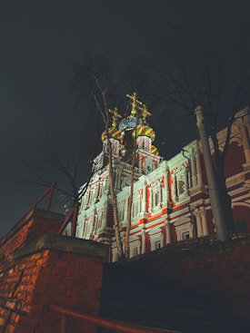 A beautifully illuminated Orthodox church with ornate architecture featuring multiple colorful domes, set against a dark, night sky. Bare trees surround the structure, adding an intriguing silhouette, while warm lights highlight the intricate designs and vibrant colors of the building.