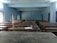 A classroom with multiple rows of wooden desks and benches arranged neatly. The room is empty and has a few ceiling fans attached to the light blue ceiling. Windows with metal bars allow natural light to enter, creating a calm atmosphere. An abandoned water bottle, notebook, and bag are on one of the desks.
