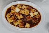 A white oval plate filled with tofu cubes covered in a dark reddish-brown sauce containing visible spices and finely chopped ingredients.