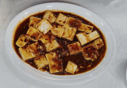 A white oval plate filled with tofu cubes covered in a dark reddish-brown sauce containing visible spices and finely chopped ingredients.