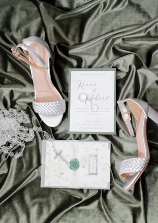 A pair of elegant women's heels with silver braided straps placed on a luxurious green satin fabric. Beside the shoes are a wedding invitation card reading 'Kiara & Robert,' accompanied by a rosary and another piece of paper with floral designs and a decorative green wax seal.