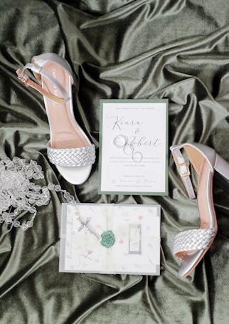 A pair of elegant women's heels with silver braided straps placed on a luxurious green satin fabric. Beside the shoes are a wedding invitation card reading 'Kiara & Robert,' accompanied by a rosary and another piece of paper with floral designs and a decorative green wax seal.