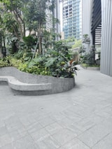 A modern architectural environment with a small garden featuring lush green plants and trees. The background shows tall glass and steel buildings, while the foreground has a curved concrete bench.
