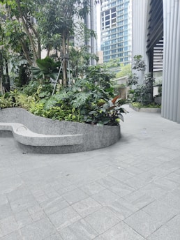 A modern architectural environment with a small garden featuring lush green plants and trees. The background shows tall glass and steel buildings, while the foreground has a curved concrete bench.