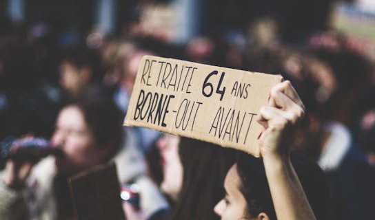 A crowd of people is gathered during a protest. One person in the foreground holds up a cardboard sign with French writing on it, indicating a political message. The focus is on the sign, while the crowd in the background is blurred.