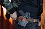 A person dressed in a camouflage jacket is wearing a gun holster on their hip. The holster contains a handgun and is attached to a brown leather belt. The background features a blurred, rusty orange surface.