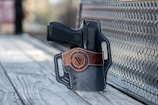 A handgun is placed inside a leather holster with brown accents. The holster rests on a textured metal surface and wooden planks, with a blurred background to the left.