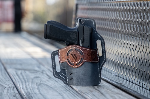 A handgun is placed inside a leather holster with brown accents. The holster rests on a textured metal surface and wooden planks, with a blurred background to the left.