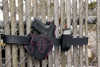 A black gun with a pink cross design on the holster, mounted on a black belt against a wooden fence with chain links.