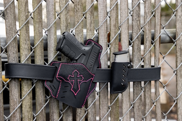 A black gun with a pink cross design on the holster, mounted on a black belt against a wooden fence with chain links.