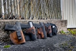 Four black handguns with brown leather holsters are lined up against a chain-link fence. The background consists of a weathered concrete wall and a wooden structure, with some greenery and leaves on the ground.