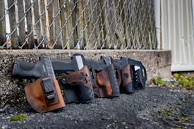 Four black handguns with brown leather holsters are lined up against a chain-link fence. The background consists of a weathered concrete wall and a wooden structure, with some greenery and leaves on the ground.