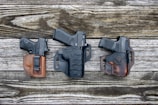 Three handguns are placed on a rustic wooden surface, each secured in its own holster. The holsters are made of leather, with two in shades of brown and one in black, showcasing detailed stitching. The arrangement is evenly spaced, showcasing the craftsmanship of the holsters.
