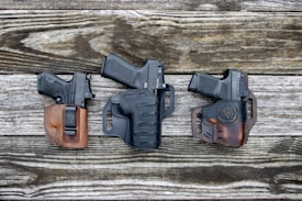 Three handguns are placed on a rustic wooden surface, each secured in its own holster. The holsters are made of leather, with two in shades of brown and one in black, showcasing detailed stitching. The arrangement is evenly spaced, showcasing the craftsmanship of the holsters.
