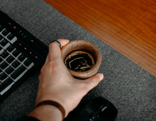 Close-up of hands typing on a keyboard with a cup of coffee beside them on a wooden desk.