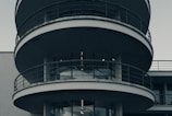 A modern circular building with multiple levels featuring curved balconies with metal railings. The structure predominantly consists of concrete and glass elements, creating a sleek and minimalistic look.