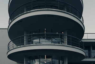A modern circular building with multiple levels featuring curved balconies with metal railings. The structure predominantly consists of concrete and glass elements, creating a sleek and minimalistic look.