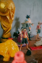 A collection of figurines is displayed on a wooden surface. A prominent golden trophy resembling the World Cup is positioned on the left, alongside various other colorful character figurines, including an anime character in a red shirt and black pants. The background is blurred, showcasing additional toys and figurines.