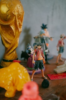 A collection of figurines is displayed on a wooden surface. A prominent golden trophy resembling the World Cup is positioned on the left, alongside various other colorful character figurines, including an anime character in a red shirt and black pants. The background is blurred, showcasing additional toys and figurines.