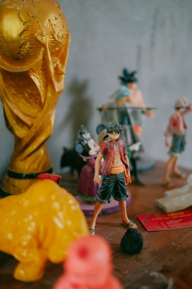 A collection of figurines is displayed on a wooden surface. A prominent golden trophy resembling the World Cup is positioned on the left, alongside various other colorful character figurines, including an anime character in a red shirt and black pants. The background is blurred, showcasing additional toys and figurines.