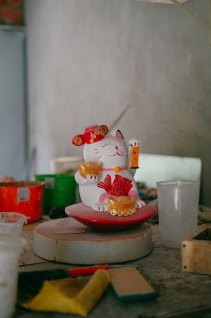 A ceramic figurine of a waving cat with traditional Chinese coin motifs is positioned on a circular platform. The cat holds a red fish and a gold ingot. Various craft materials and containers are scattered around on a dusty worktable, suggesting an artistic workshop environment.