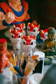 A collection of decorative items including several small statues resembling dragons or lions with red and gold features, a golden Buddha statue, art supplies such as paintbrushes, and part of an action figure in an orange and blue outfit.