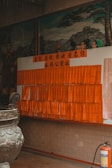 A display of numerous orange tags with Chinese characters is mounted on a wall in a dimly lit space. In front of the display, there is a large, ornate incense burner with intricate patterns. The scene includes a fire extinguisher in the right corner and murals of traditional Chinese figures and landscapes in the background.