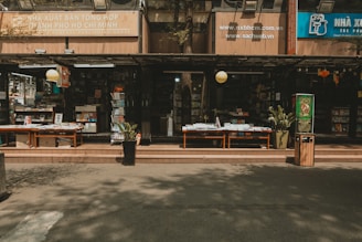 An outdoor bookstore with multiple shelves stacked with a variety of books. There are tables displaying books under a shaded awning, with potted plants and a trash bin nearby. The signage indicates it is in Ho Chi Minh City, and there is a calm and quiet atmosphere.