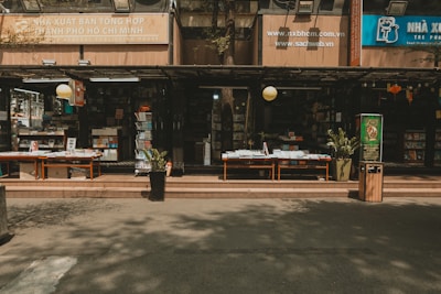 An outdoor bookstore with multiple shelves stacked with a variety of books. There are tables displaying books under a shaded awning, with potted plants and a trash bin nearby. The signage indicates it is in Ho Chi Minh City, and there is a calm and quiet atmosphere.