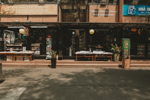 An outdoor bookstore with multiple shelves stacked with a variety of books. There are tables displaying books under a shaded awning, with potted plants and a trash bin nearby. The signage indicates it is in Ho Chi Minh City, and there is a calm and quiet atmosphere.
