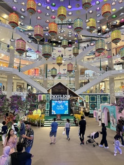 A vibrant shopping mall interior features numerous decorative lanterns suspended from the ceiling. The central area displays a traditional decorative structure with a sign labeled Pavilion Kuala Lumpur. Shoppers are seen walking around and engaging in activities on multiple levels connected by escalators and stairs.
