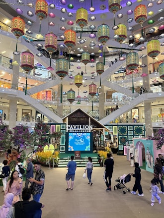 A vibrant shopping mall interior features numerous decorative lanterns suspended from the ceiling. The central area displays a traditional decorative structure with a sign labeled Pavilion Kuala Lumpur. Shoppers are seen walking around and engaging in activities on multiple levels connected by escalators and stairs.