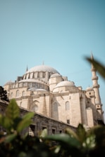 An impressive historical mosque with a large central dome and multiple smaller domes, displaying classic Islamic architecture. The structure is made of light-colored stone, and a tall minaret is visible in the background. In the foreground, blurred foliage adds depth to the image.