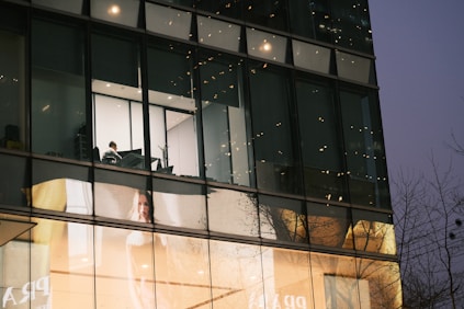 A modern building exterior features large glass windows, reflecting the surrounding environment. Inside, a solitary person is visible working at a desk in a brightly lit room. Below, a large advertisement displays a woman against a softly lit background.