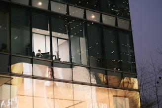 A modern building exterior features large glass windows, reflecting the surrounding environment. Inside, a solitary person is visible working at a desk in a brightly lit room. Below, a large advertisement displays a woman against a softly lit background.