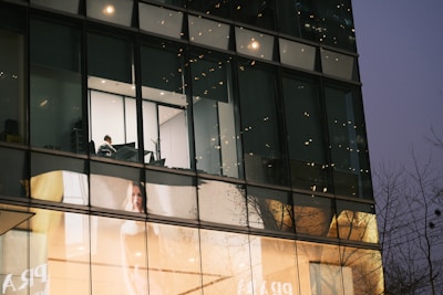 A modern building exterior features large glass windows, reflecting the surrounding environment. Inside, a solitary person is visible working at a desk in a brightly lit room. Below, a large advertisement displays a woman against a softly lit background.
