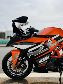 A sleek and sporty orange and black motorcycle is parked on the side of the road. The bike features aerodynamic design elements with sharp angles and bold graphics. The brand logo is visible on the body of the motorcycle, and it is positioned against a blurred background, suggesting an urban or suburban setting.